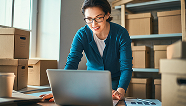 smiling business woman using laptop on desk with stacks of boxes throughout office area